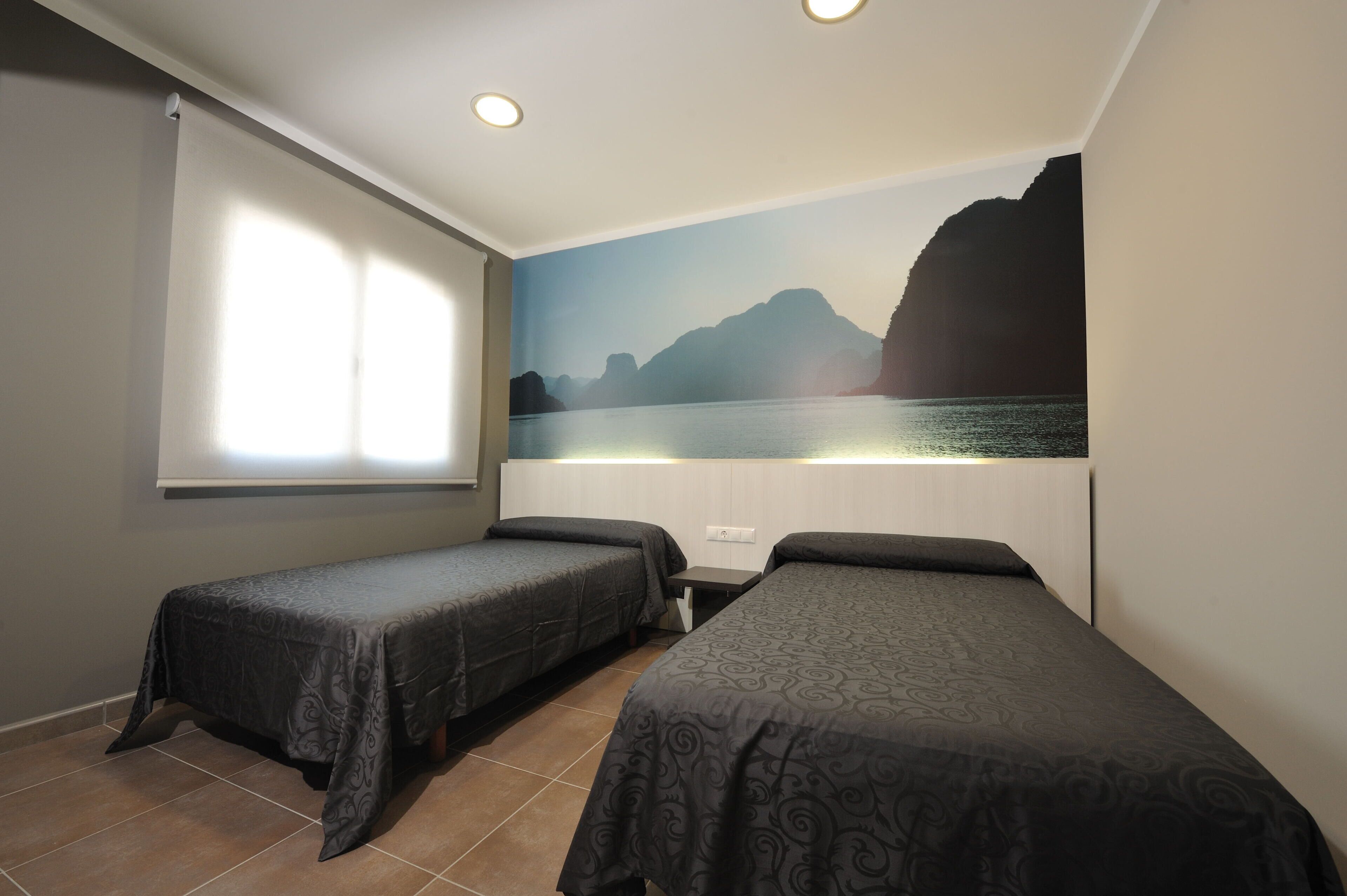Una habitación con dos camas individuales, una ventana con persianas, un mural en la pared con una escena costera y una mesita.