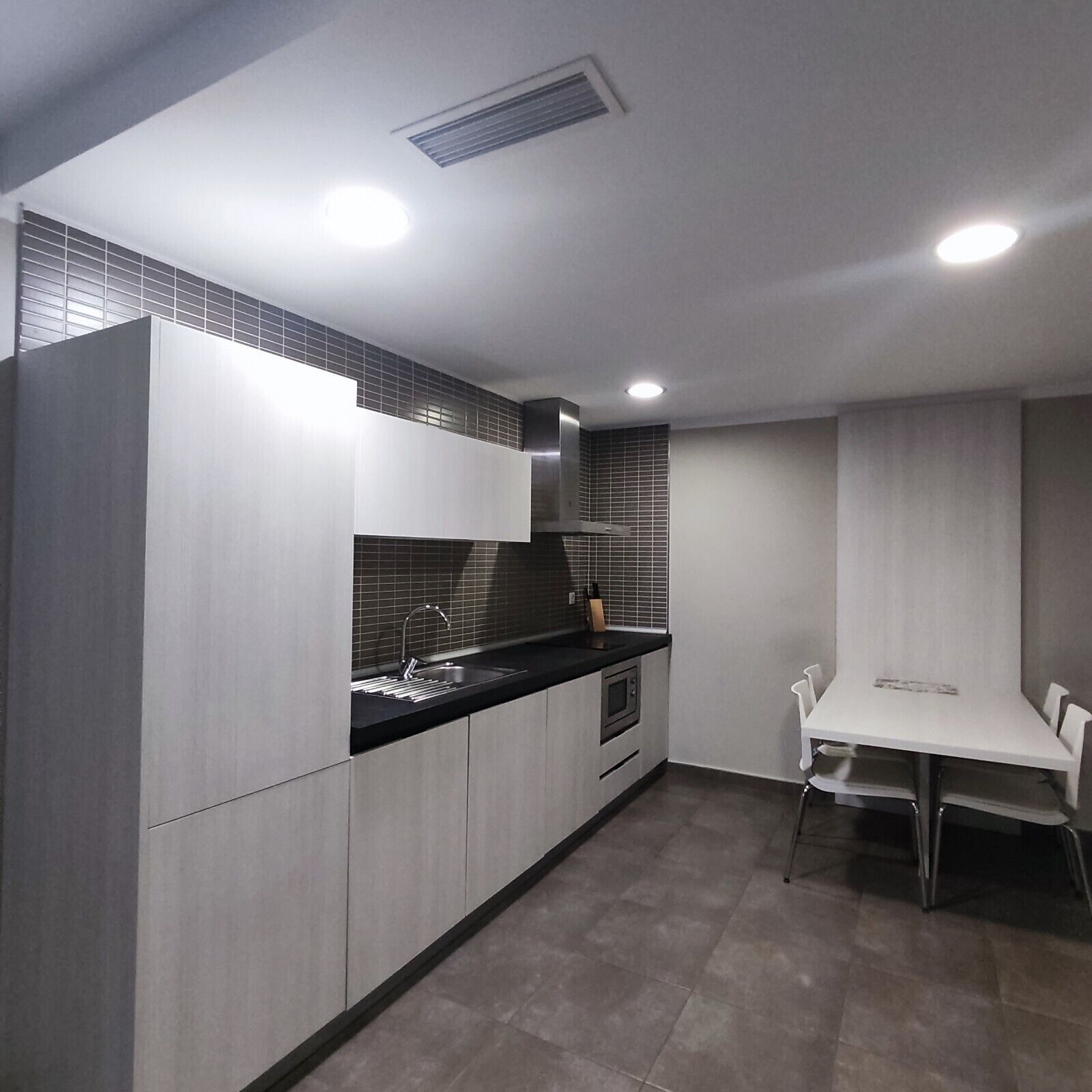 A modern kitchen with grey cabinetry, a black countertop, a built-in oven, and a dining area with a white table and chairs.