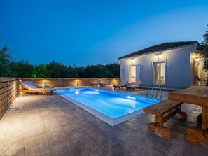 Uma piscina retangular com um deck de madeira e cadeiras de descanso, cercada por uma vedação de madeira e um pequeno edifício com telhado ladrilhado.