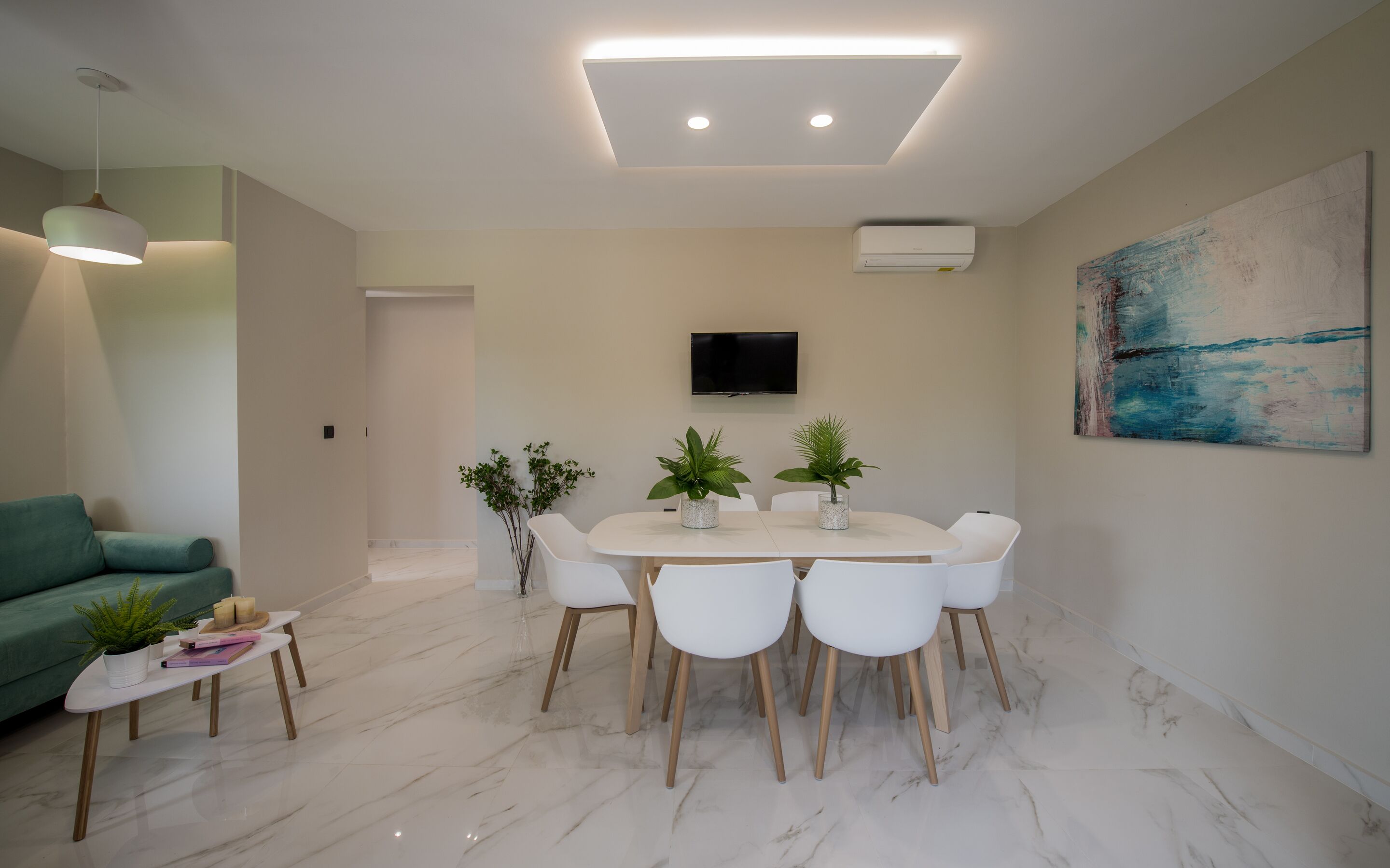 Un espace repas moderne avec une table et des chaises blanches, un luminaire au plafond et un téléviseur à écran plat fixé au mur.