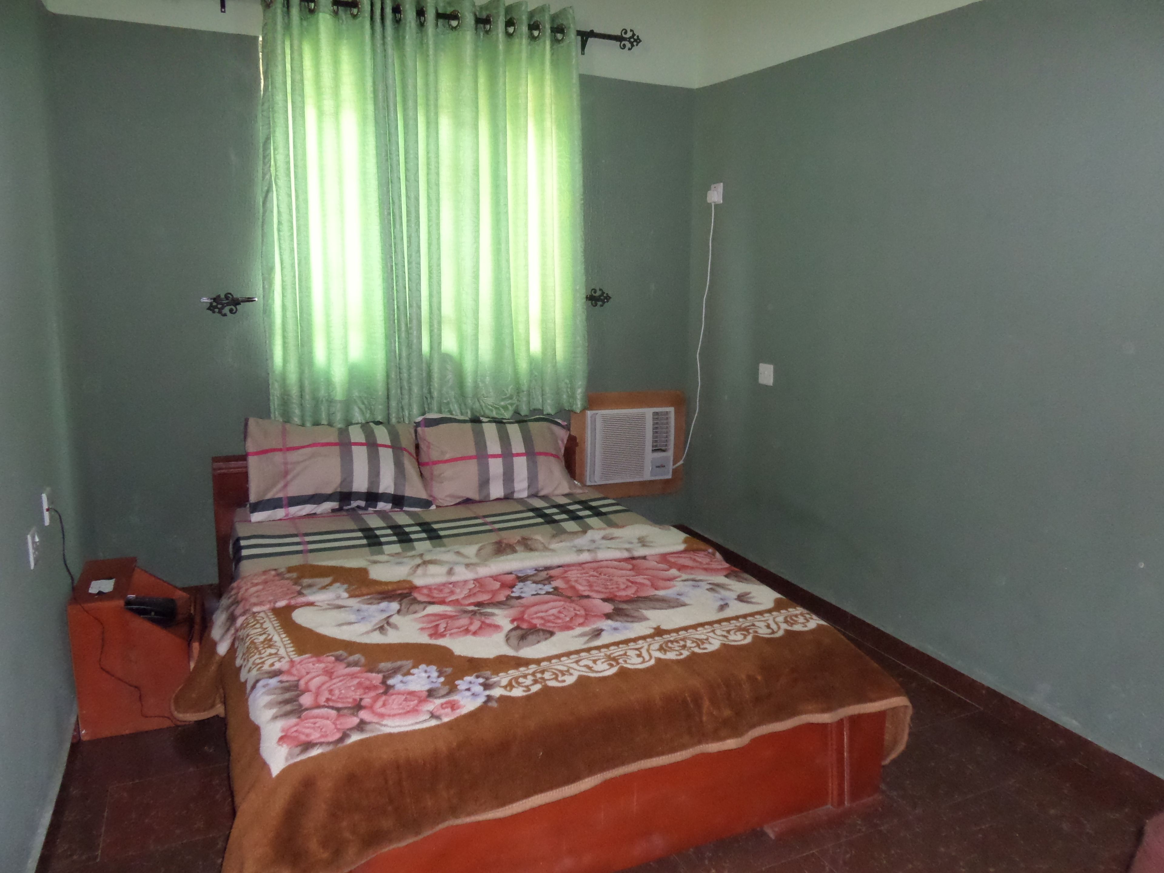 A bedroom with a bed, a window with curtains, an air conditioning unit, and a wall socket.