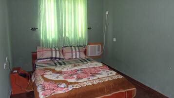 A bedroom with a bed, a window with curtains, an air conditioning unit, and a wall socket.