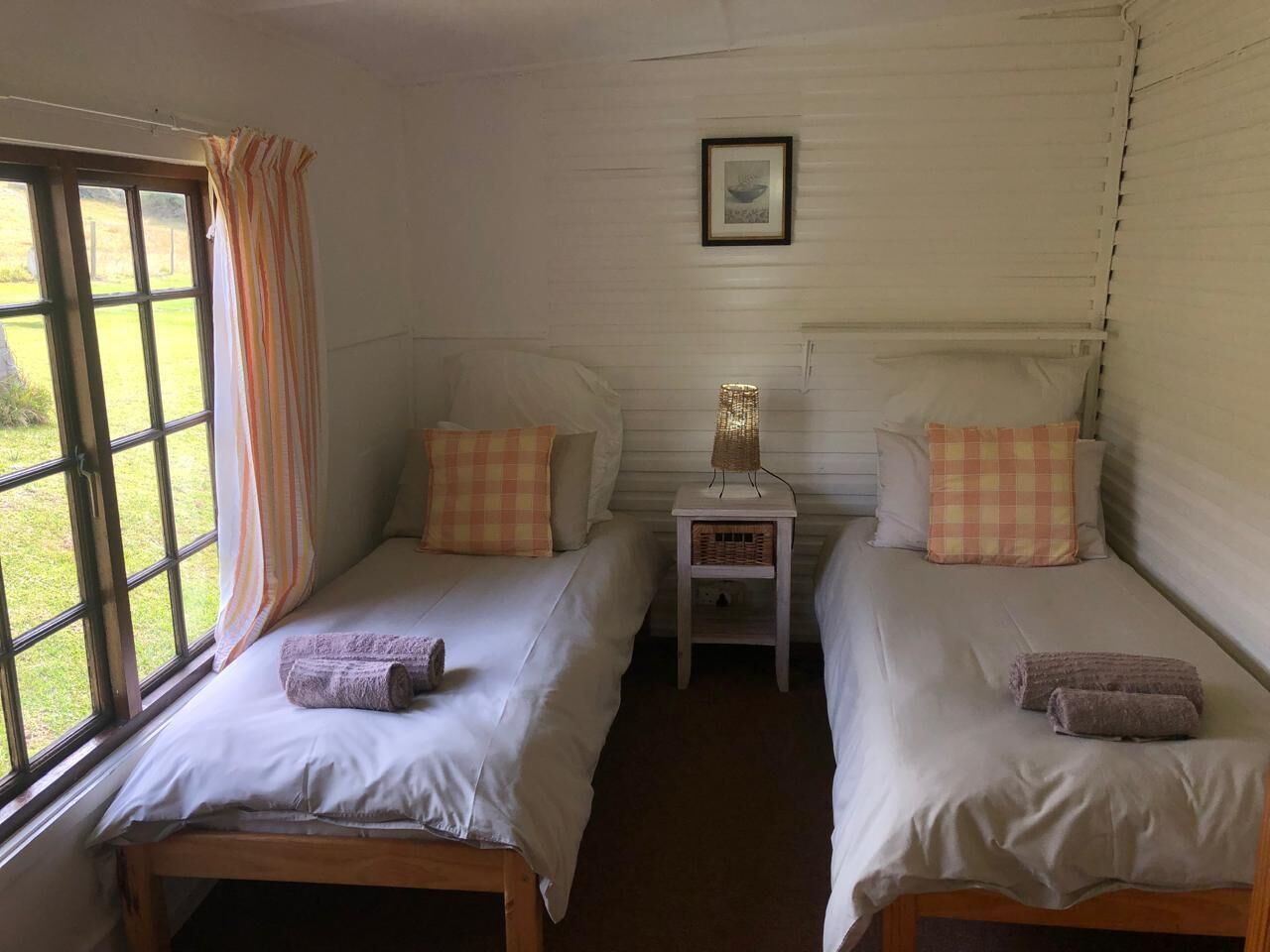 A room with two beds, a small table with a lamp, a window with curtains, and a framed picture on the wall.