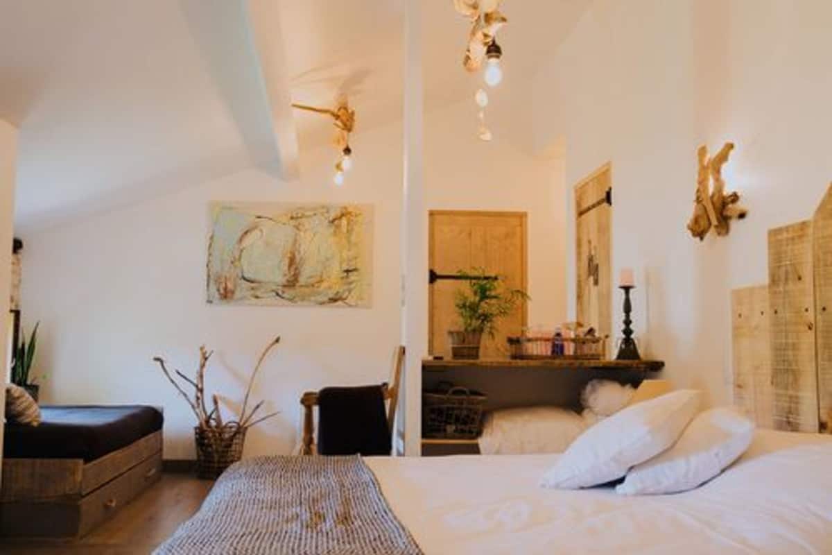 A bedroom with a bed, a chair, a small table, a wall-mounted shelf, and a ceiling fan.