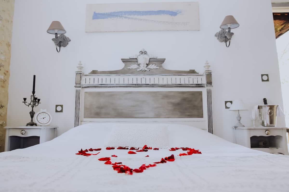 A neatly made bed with a heart-shaped arrangement of rose petals in the center, flanked by two bedside tables with lamps and a clock.