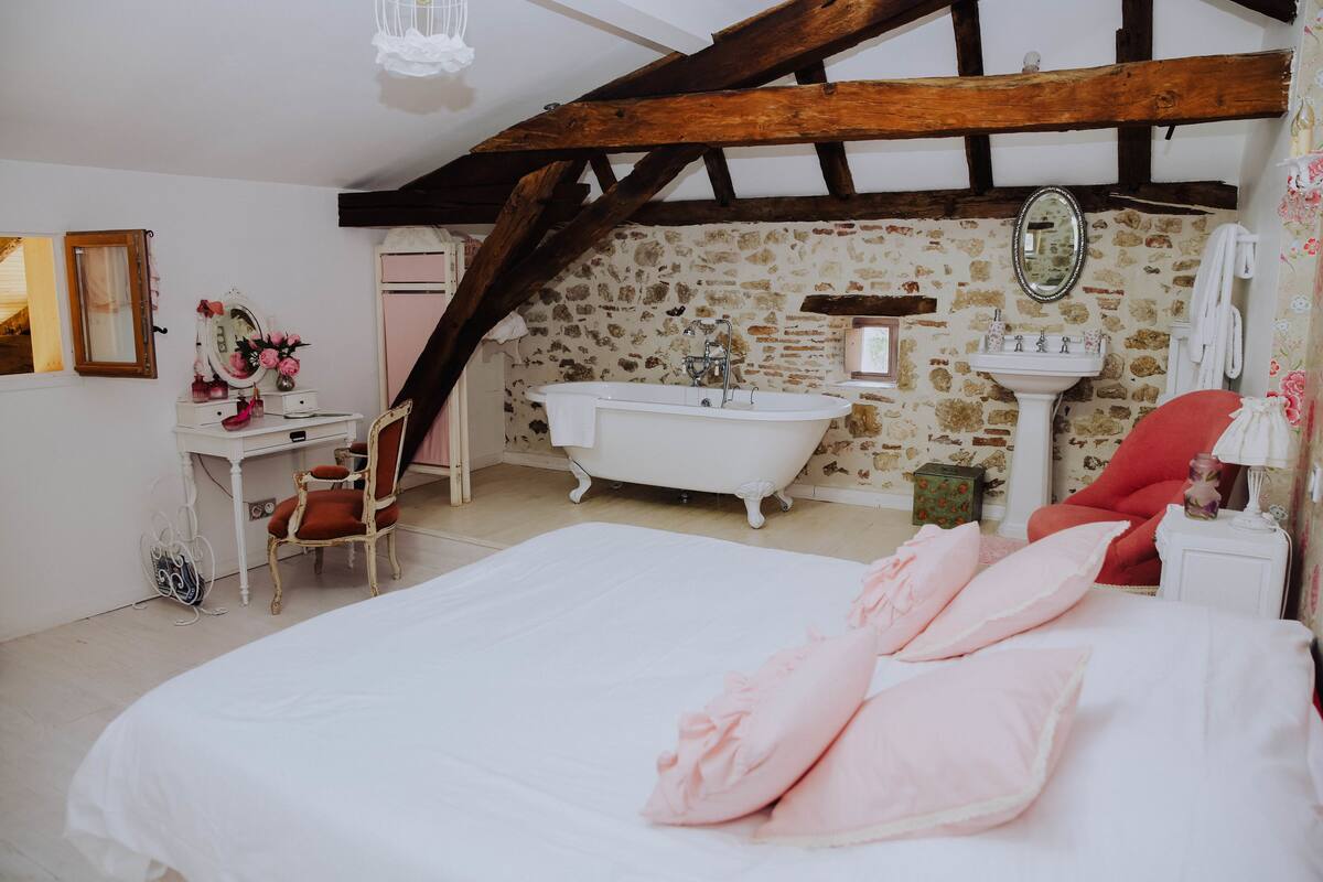 A bedroom with a large bed, a vanity with a mirror, a clawfoot bathtub, and a wooden ceiling beam.