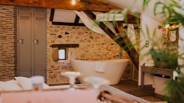 A rustic room with stone walls, a white bathtub, and a wooden ceiling. - La Maison Bleue (Villereal)