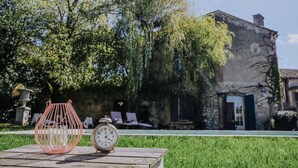 A wooden table with a red wire basket and a classic alarm clock. In the background, there is a stone building with ivy and a pool area. - La Maison Bleue (Villereal)