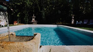 A swimming pool with a fountain, surrounded by trees and a stone structure. - La Maison Bleue (Villereal)