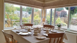 A dining area with a table set for four, large windows, and a view of a garden and buildings outside. - Shian Cottage B&B (Dunkeld)