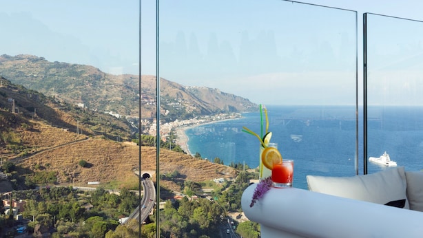 Une pièce dotée d’une grande fenêtre donnant sur un paysage côtier, avec une boisson sur la table et une vue sur un pont et des collines.