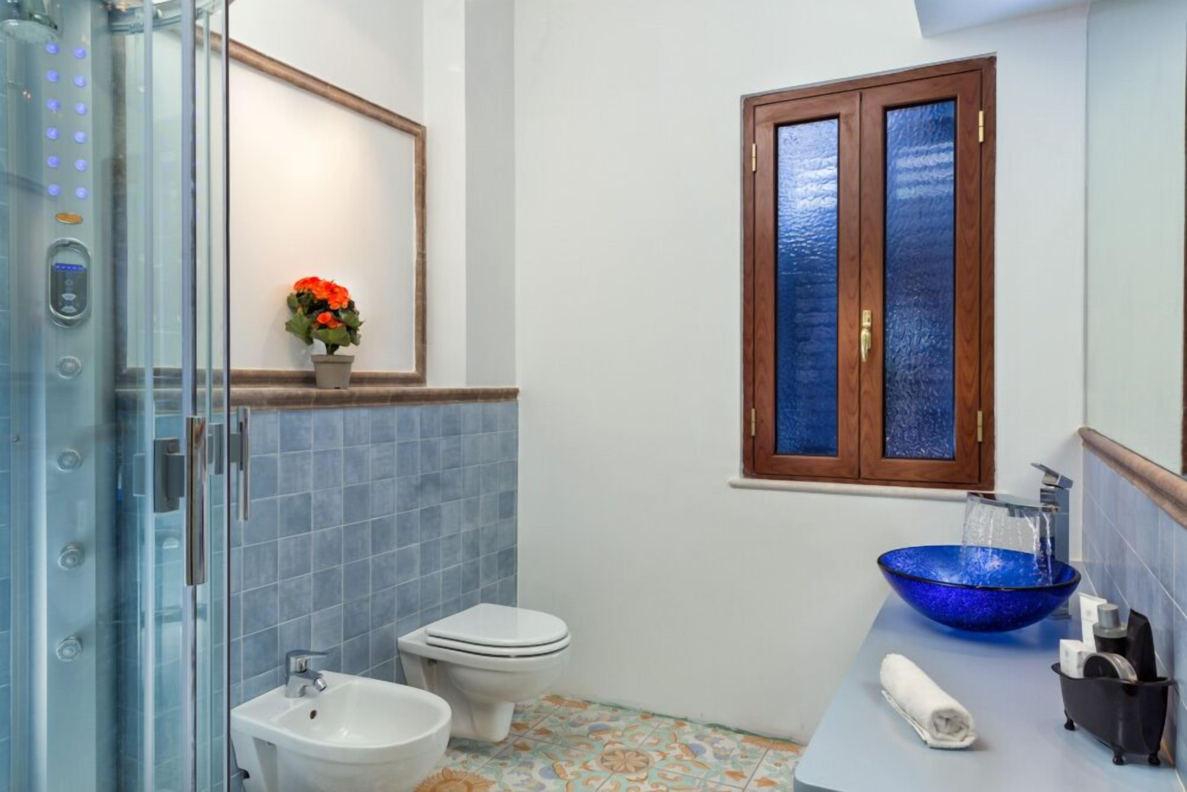 Ein Badezimmer mit Duschkabine aus Glas, einer weißen Toilette, einem blauen Glaswaschbecken und einem Holzschrank mit Fenster darüber.