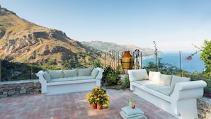 Une terrasse dotée de sièges blancs et confortables, agrémentée d’un pot de fleurs, qui offre une vue imprenable sur un paysage montagneux et la mer.