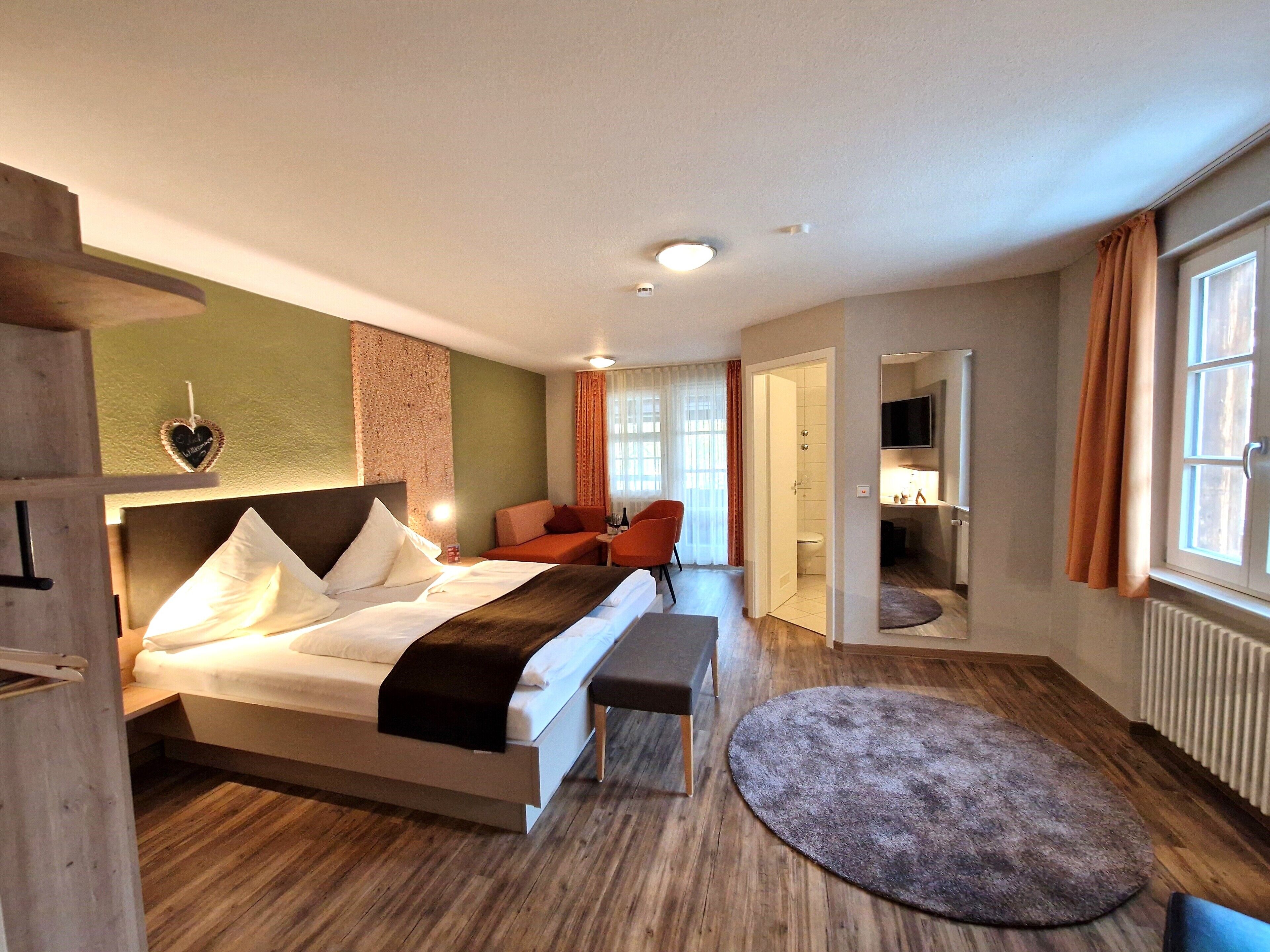 A hotel room with a bed, a bench, a small table, and a bathroom visible through an open door.
