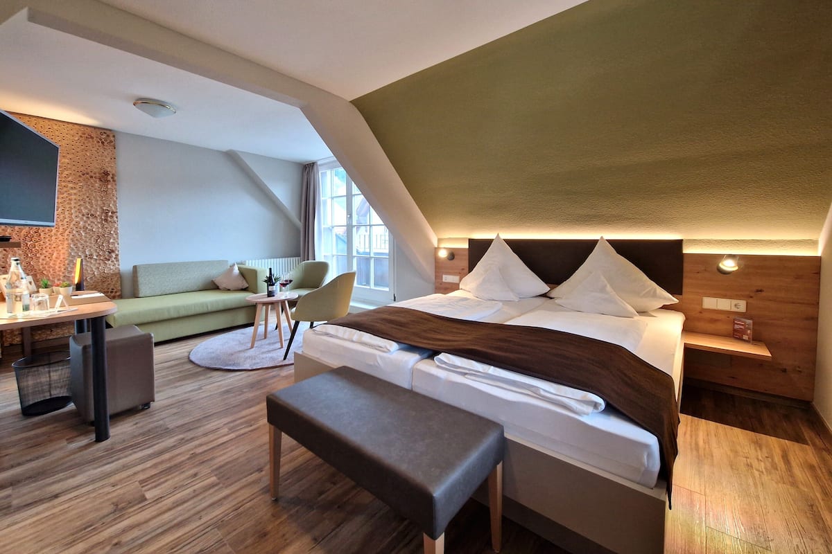 Ein modernes, helles Zimmer mit einem großen Bett, einer Sitzecke mit Sofa und Sesseln, einem kleinen Tisch mit Getränken und einem Fernseher.