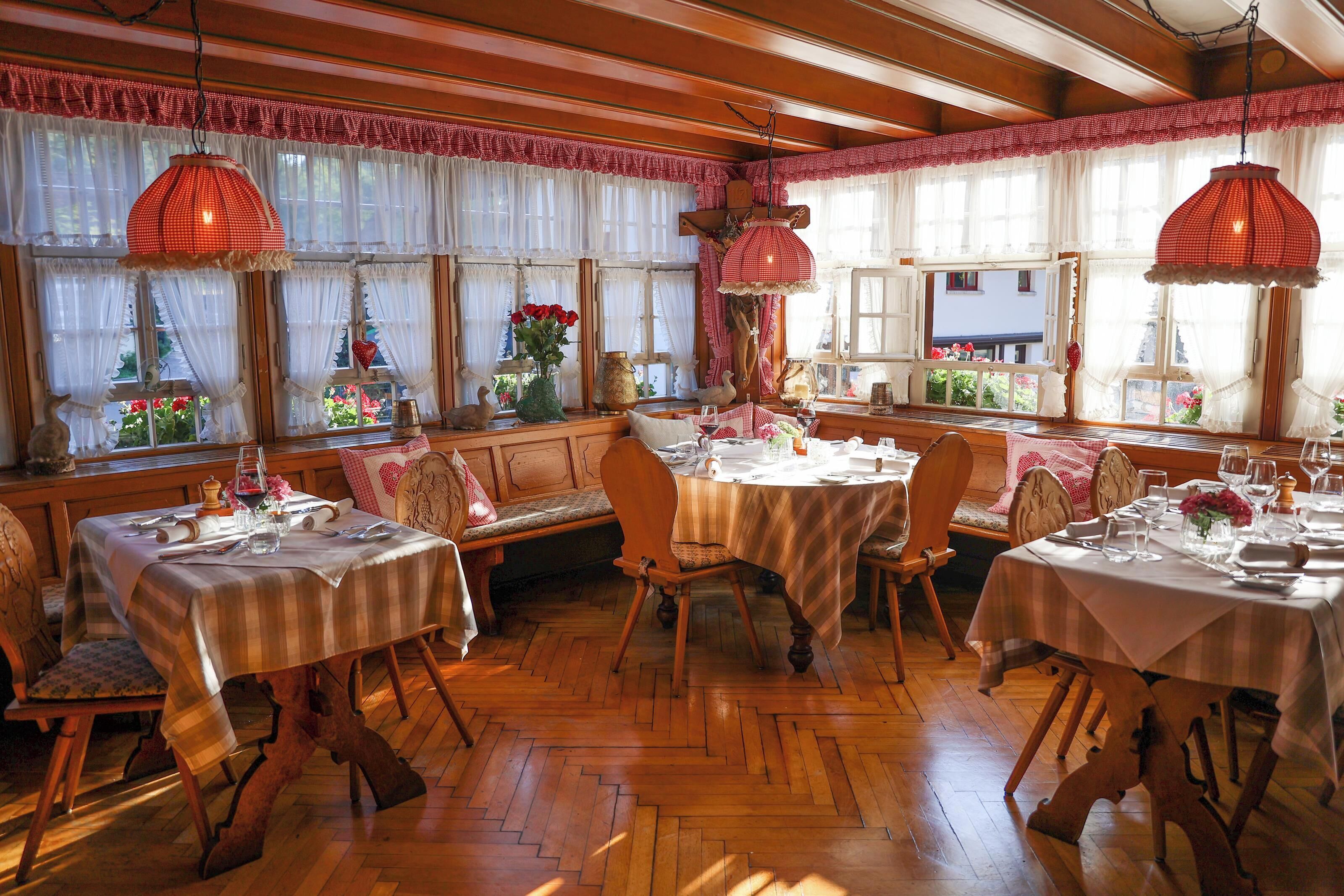 Zona pranzo con tavoli e sedie in legno, ampie finestre con tende e lampade rosse pendenti.