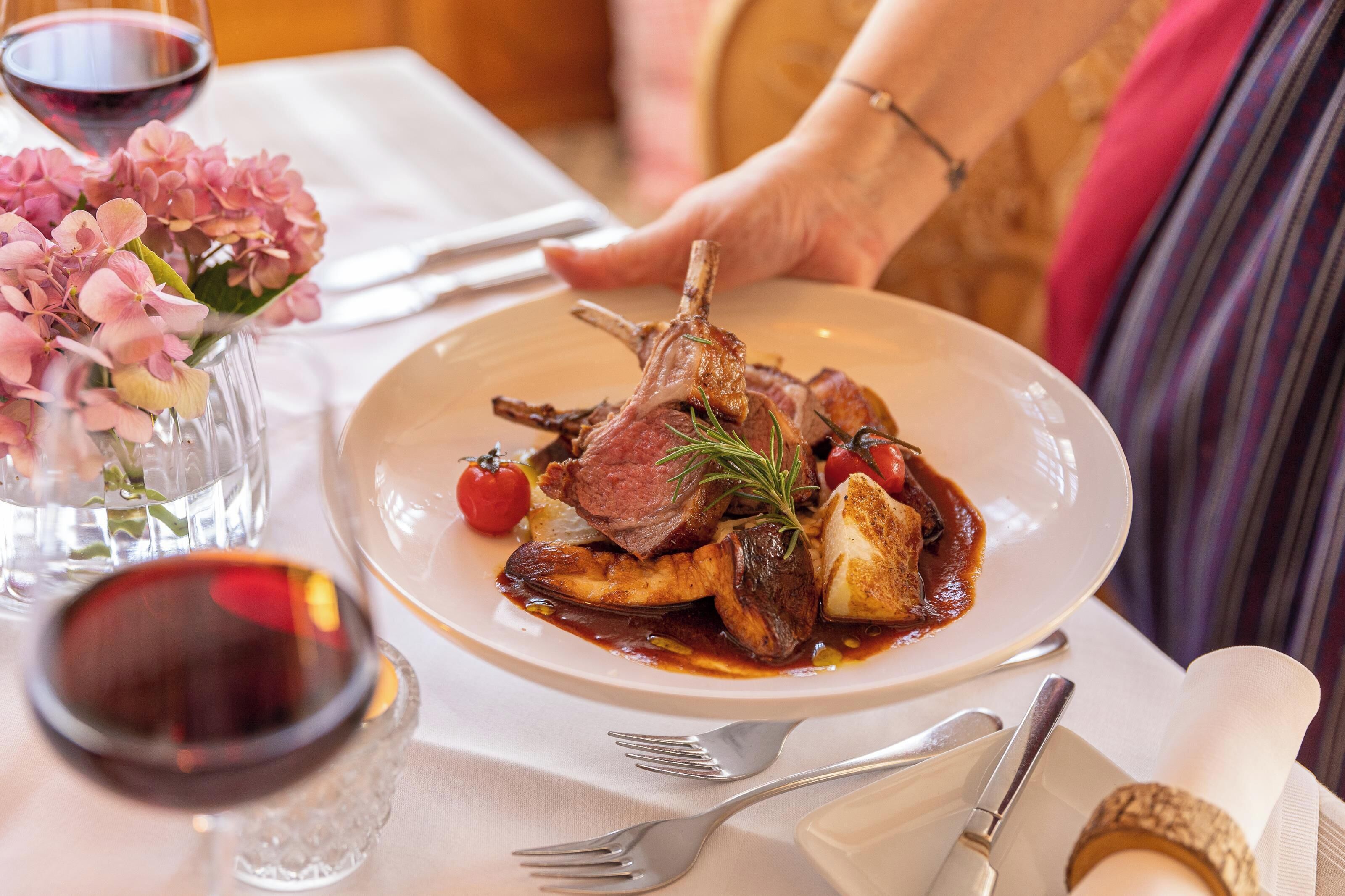 A plate of food with a lamb shank, roasted potatoes, and cherry tomatoes, accompanied by a glass of red wine and a vase of pink flowers.