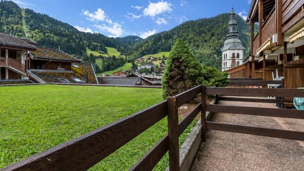 A mountain village with traditional wooden buildings, a church with a bell tower, and a clear view of the surrounding hills. - 3-room apartment, terrace, pets allowed, garage, village center (La Clusaz)