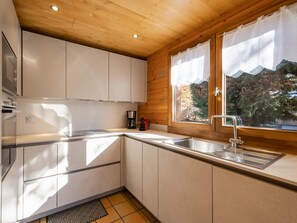 A modern kitchen with wooden cabinets, a stainless steel sink, and a window with a view of trees. - 3* Chalet for 10, fireplace, garage, WIFI (La Clusaz)