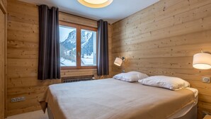 A wooden-paneled room with a bed, a window with curtains, a wall-mounted lamp, and a radiator.