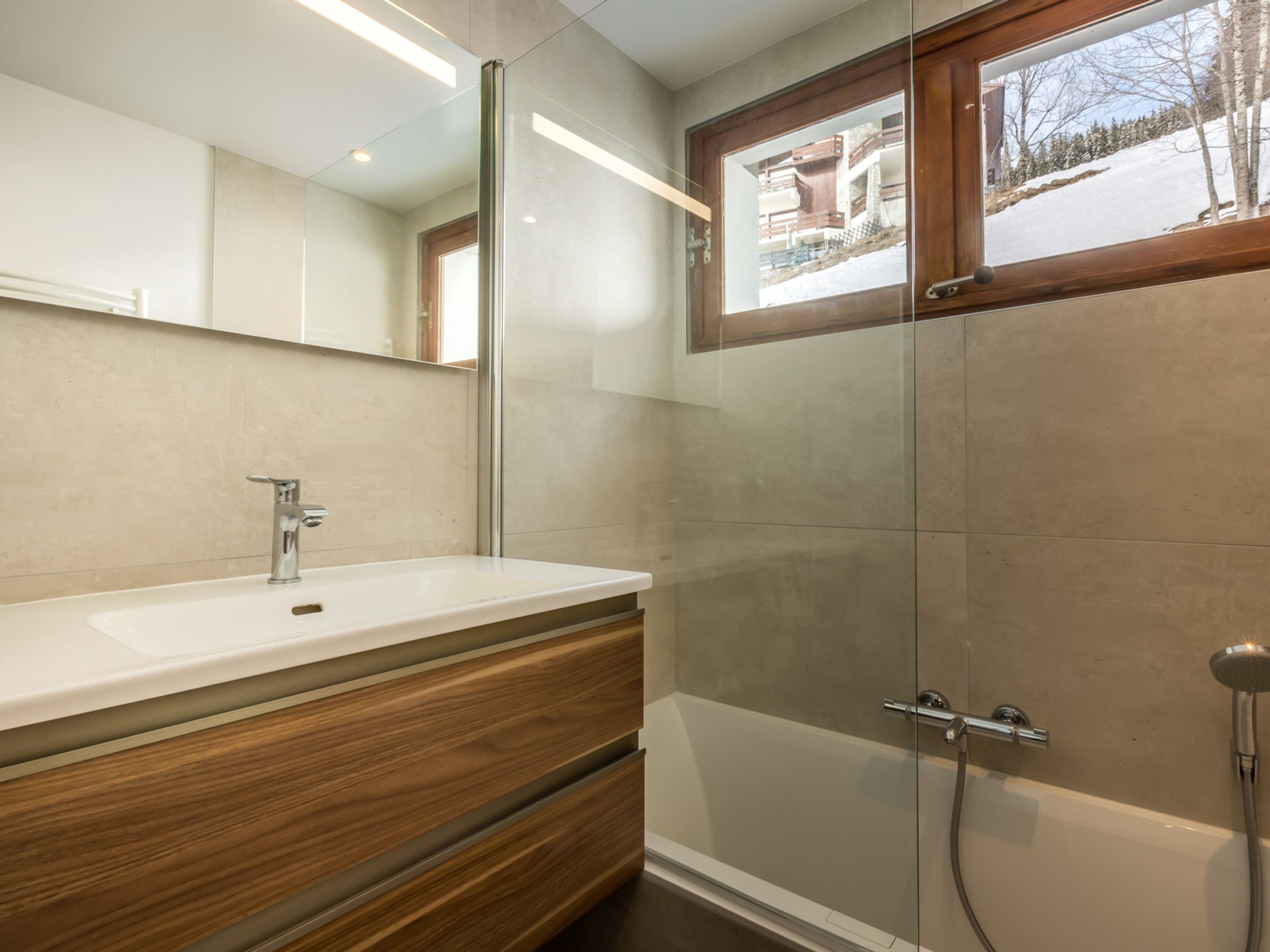 A modern bathroom with a large mirror, a white sink, wooden cabinetry, a glass shower enclosure, and a view of a snowy landscape through a window.