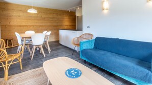 A modern living room with a blue sofa, wooden dining table with white chairs, a wooden accent wall, and a kitchen area.