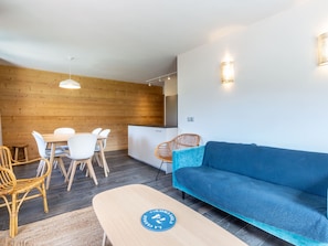 A modern living room with a blue sofa, wooden dining table with white chairs, a wooden accent wall, and a kitchen area.