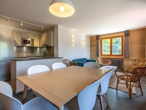 Une cuisine moderne équipée d’un four à micro-ondes et d’une cuisinière, un coin repas avec une table en bois et des chaises blanches, et une fenêtre donnant sur les arbres.