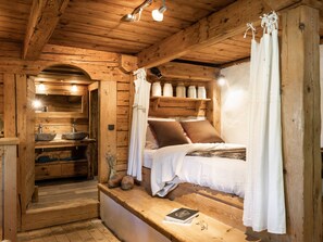 A rustic wooden cabin interior with a bed, a wooden bench with a book, a sink, and a shelf with jars.