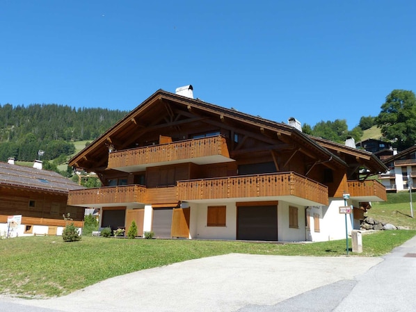 A two-story wooden chalet with balconies, a garage, and a view of a forested hillside.
