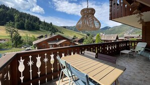 Un balcon en bois, agrémenté d’une table et de chaises, qui offre une vue sur un paysage montagneux avec des maisons et un ciel dégagé.