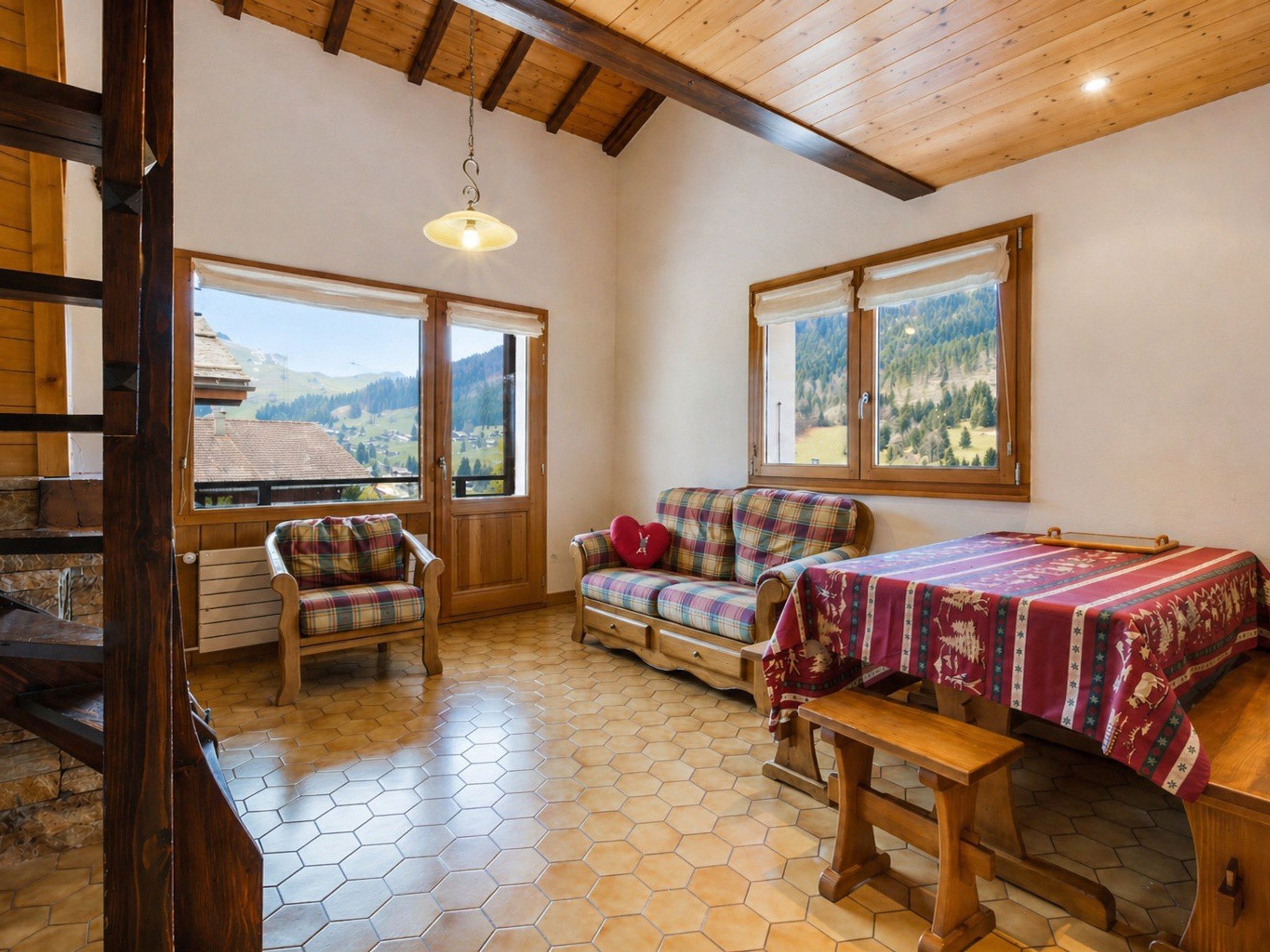A cozy room with a wooden bench, a sofa, and a table with a patterned tablecloth. Large windows offer a view of the mountains.
