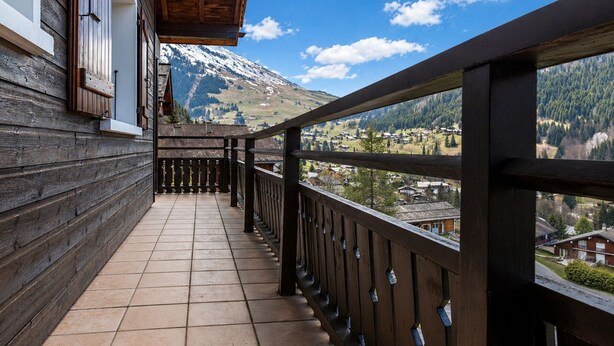 Een houten balkon met betegelde vloer, houten hekwerk en uitzicht op een berg en dorp.