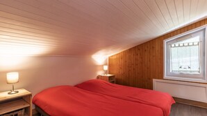 A bedroom with a wooden ceiling, a window with a curtain, a bed with red bedding, a bedside table with a lamp, and a small wooden shelf. - Spacious apt 7 ppl, 3*, quiet, La Clusaz (La Clusaz)