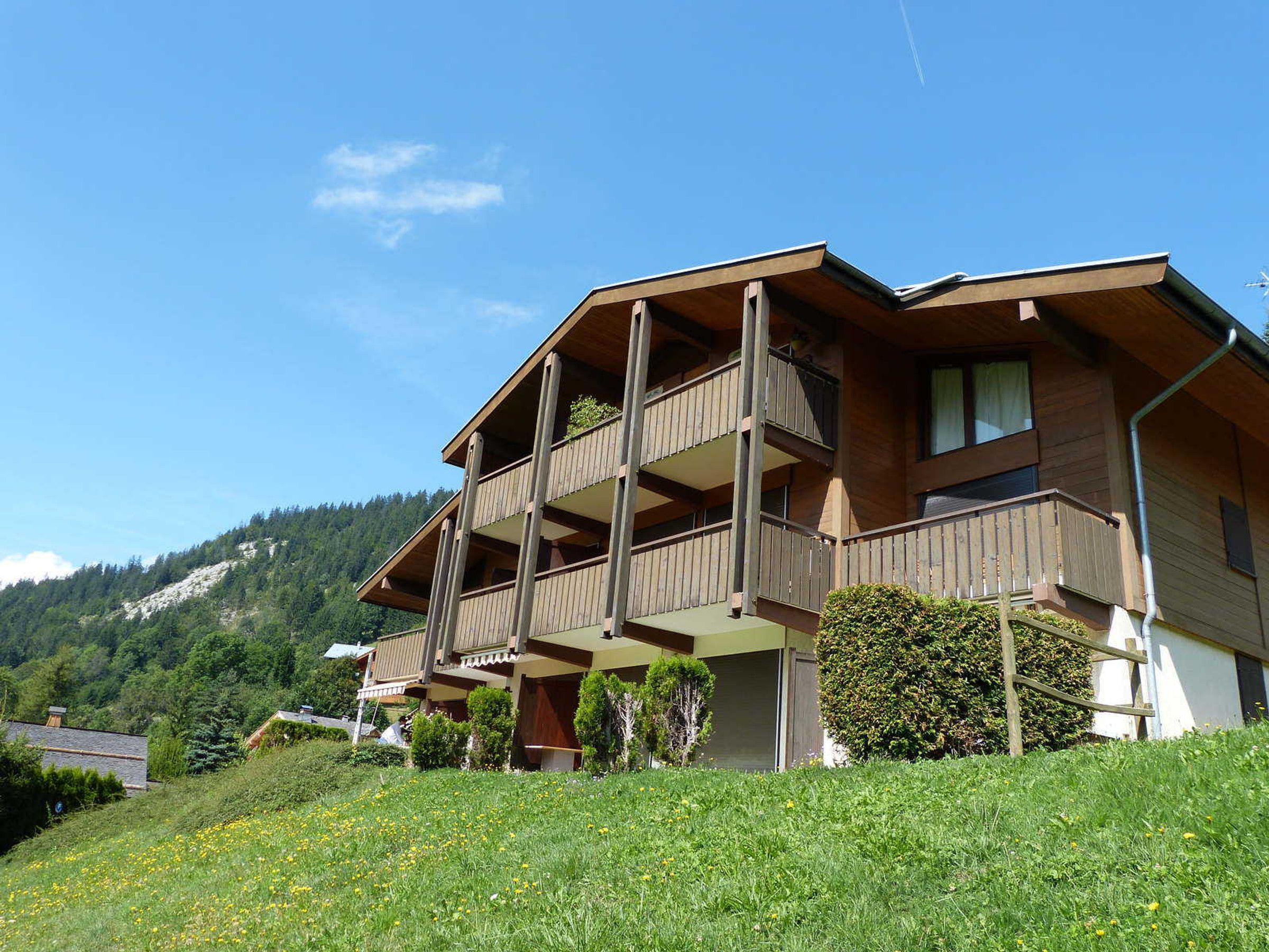 A multi-story wooden building with balconies, surrounded by a grassy area and a forested hill.