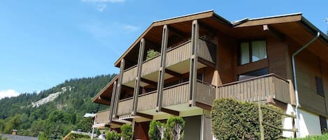 Ein mehrstöckiges Holzgebäude mit Balkonen, umgeben von einer Wiese und einem bewaldeten Hügel.