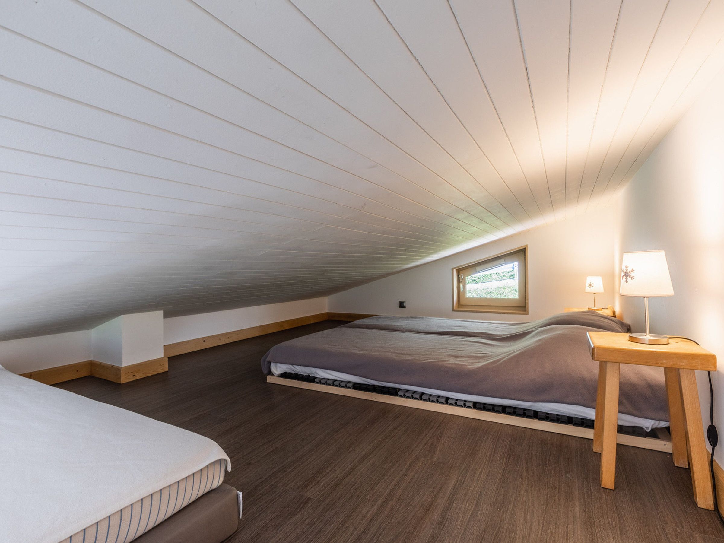 A bedroom with a sloped ceiling, a bed, a bedside table with a lamp, and a window.
