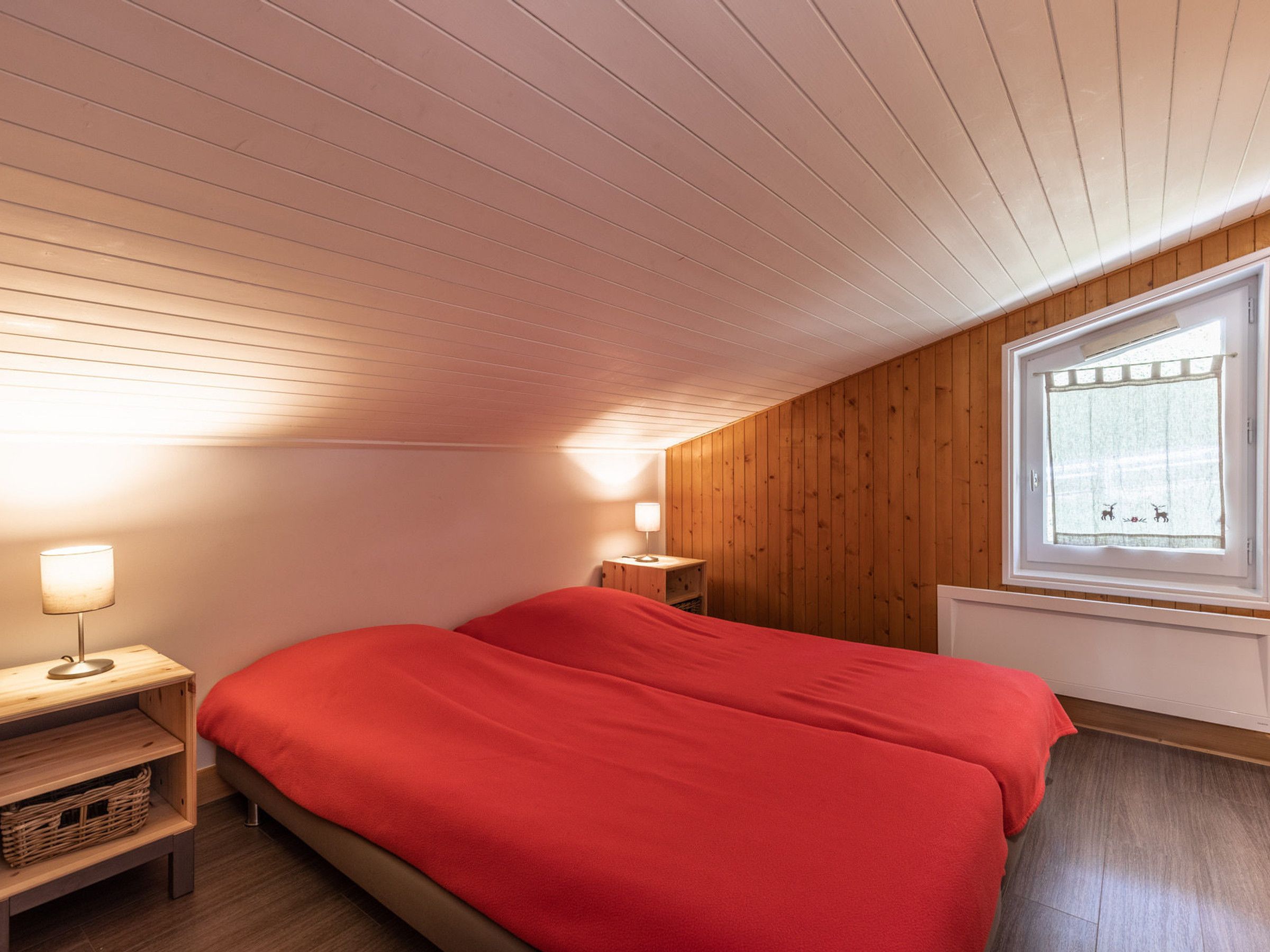 A bedroom with a wooden ceiling, a window with a curtain, a bed with red bedding, a bedside table with a lamp, and a small wooden shelf.