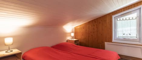A bedroom with a wooden ceiling, a window with a curtain, a bed with red bedding, a bedside table with a lamp, and a small wooden shelf.