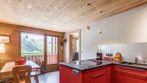 A modern kitchen with red cabinetry, a wooden dining area, and a view of a green landscape through large windows. - Spacious apt 7 ppl, 3*, quiet, La Clusaz (La Clusaz)