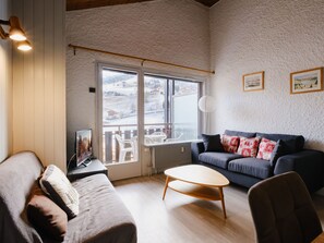 A cozy living room with a sofa, a coffee table, and a TV on a stand. There is a large window with a view of a snowy landscape. - Charming studio near slopes, pets allowed (La Clusaz)