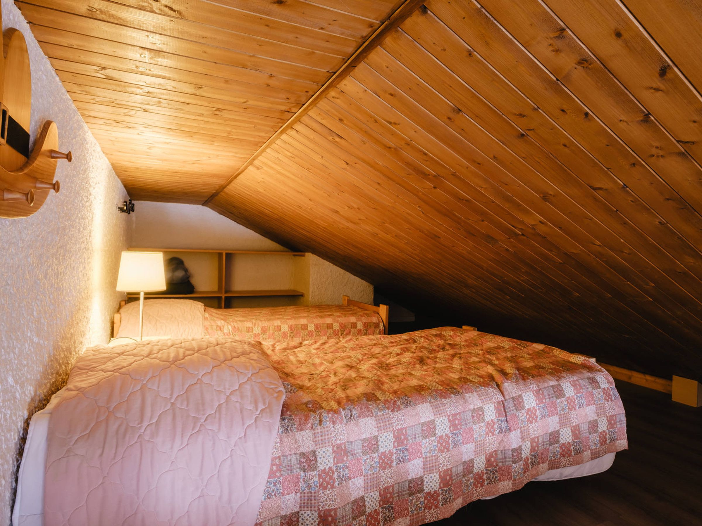 An attic bedroom with wooden beams, a bed with a quilt, a bedside table, and a lamp.