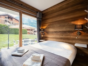 A hotel room with a large bed, a wooden headboard, a window with a view of a mountain and a building, and a table with chairs outside. - 3★ apt, 6 guests, south terr., center La Clusaz, 50m to slopes, pets allowed (La Clusaz)
