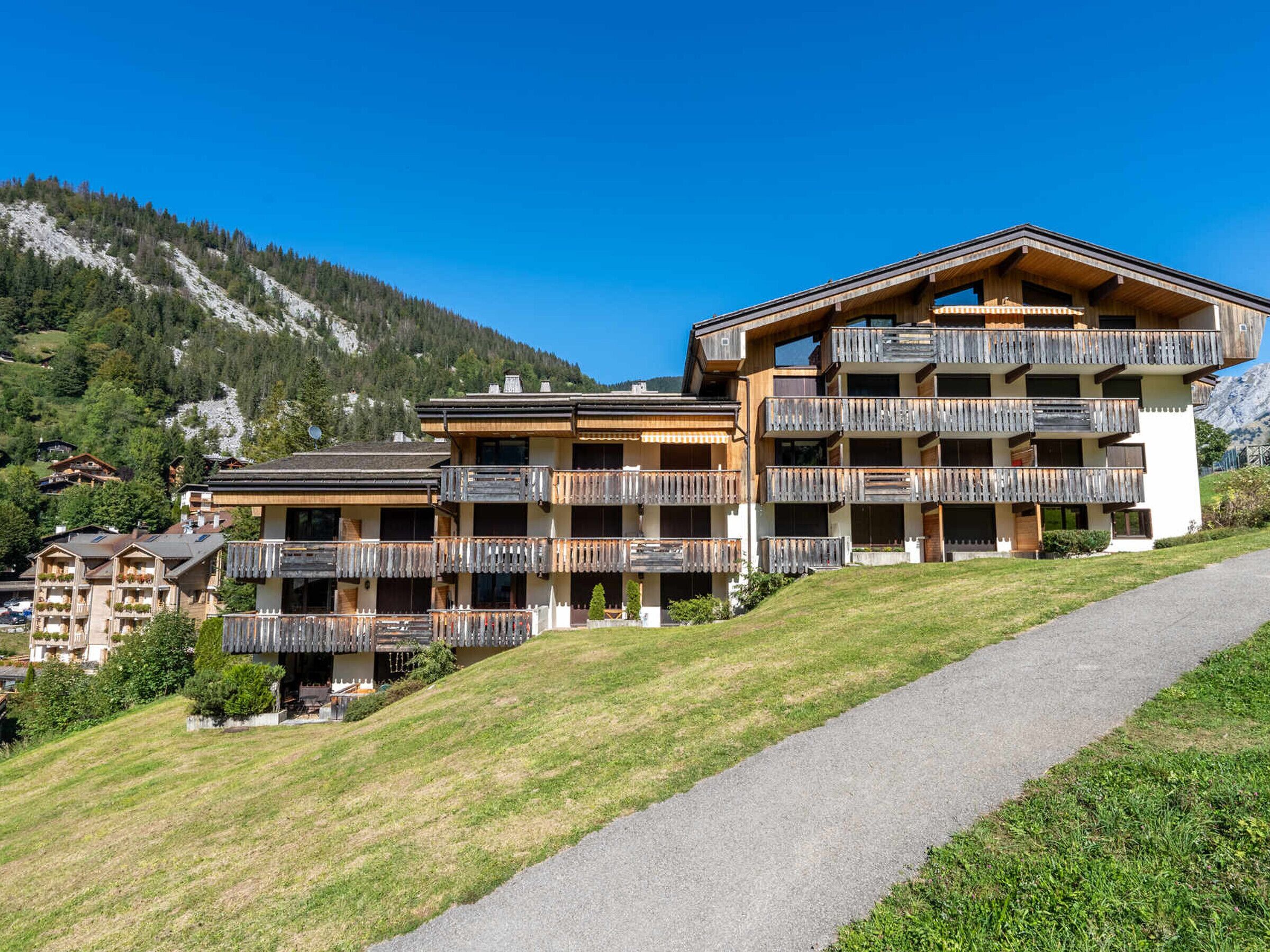 A multi-story hotel with wooden balconies, a paved pathway, and a mountainous backdrop.