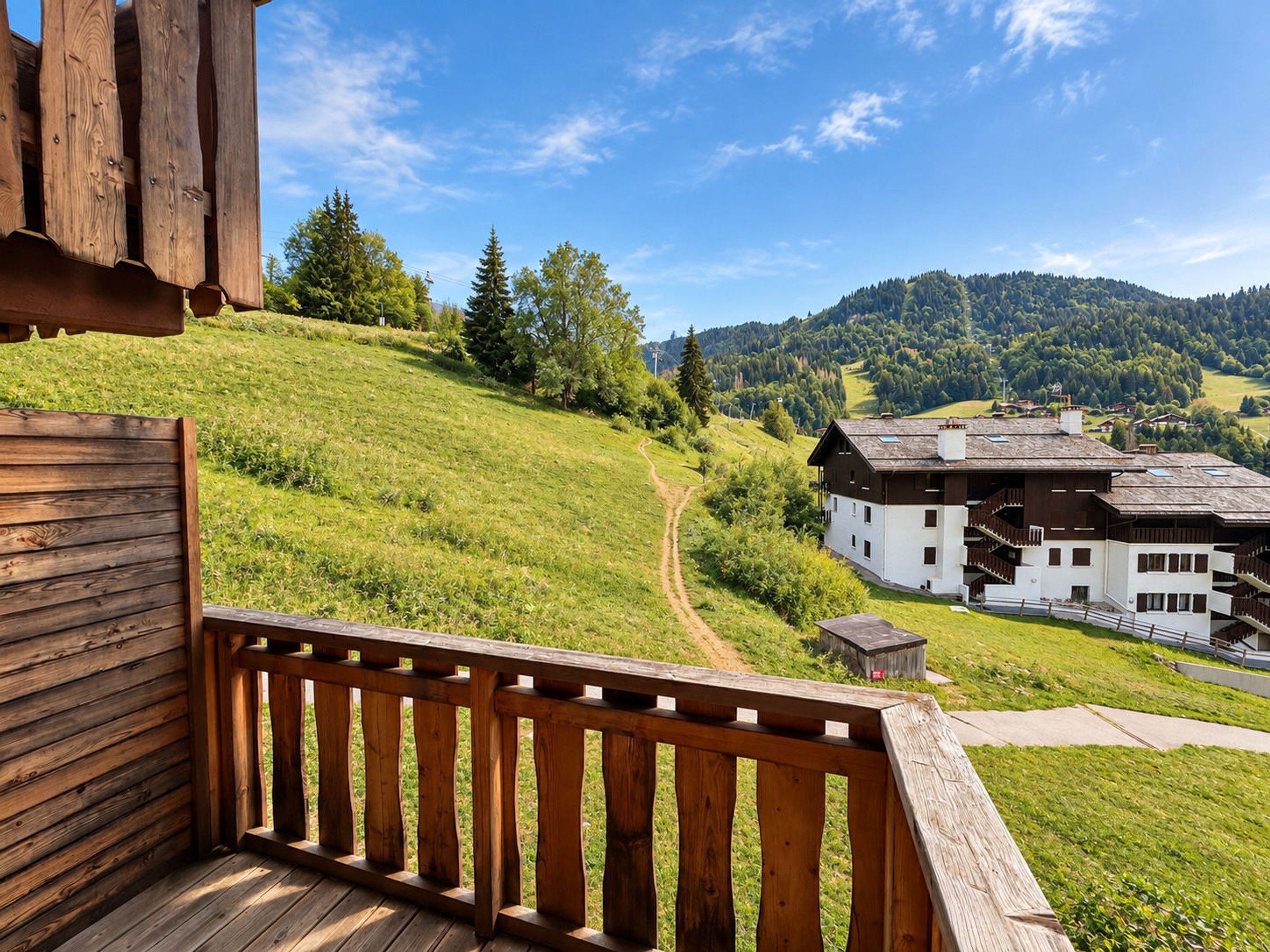 Eine Holzterrasse mit Blick auf ein weißes Gebäude mit Balkonen, umgeben von grünen Hügeln und einem klaren Himmel.