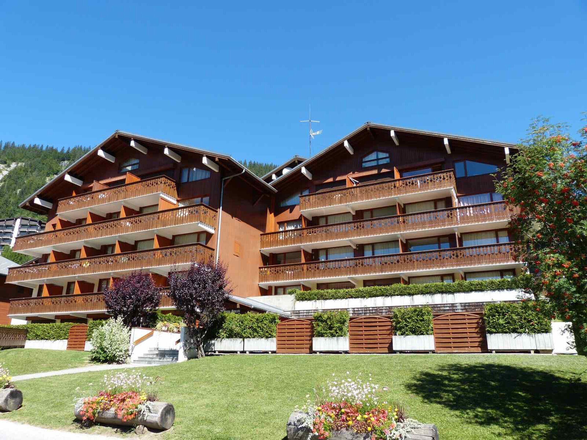 A multi-story hotel with balconies, a ski slope in the background, and well-maintained gardens.