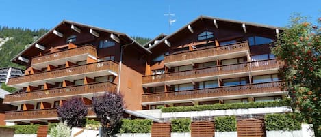 Un hotel de varios pisos con balcones, una pista de esquí al fondo y jardines cuidados.