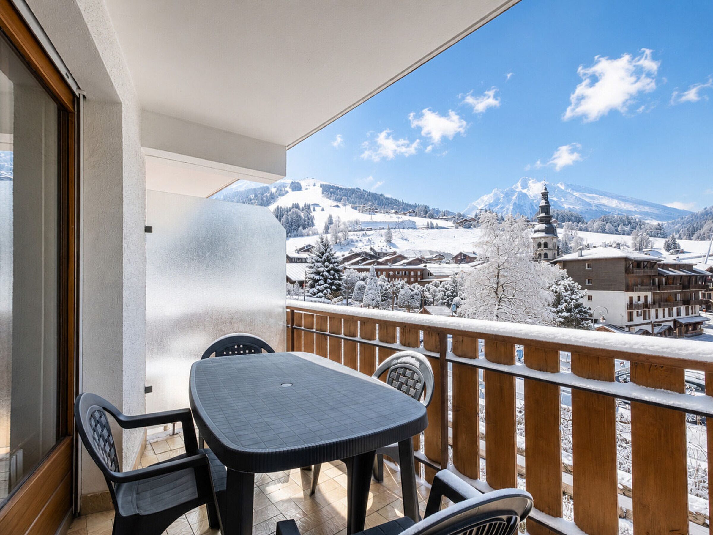 A balcony with a table and chairs, overlooking a snowy mountain landscape with buildings.