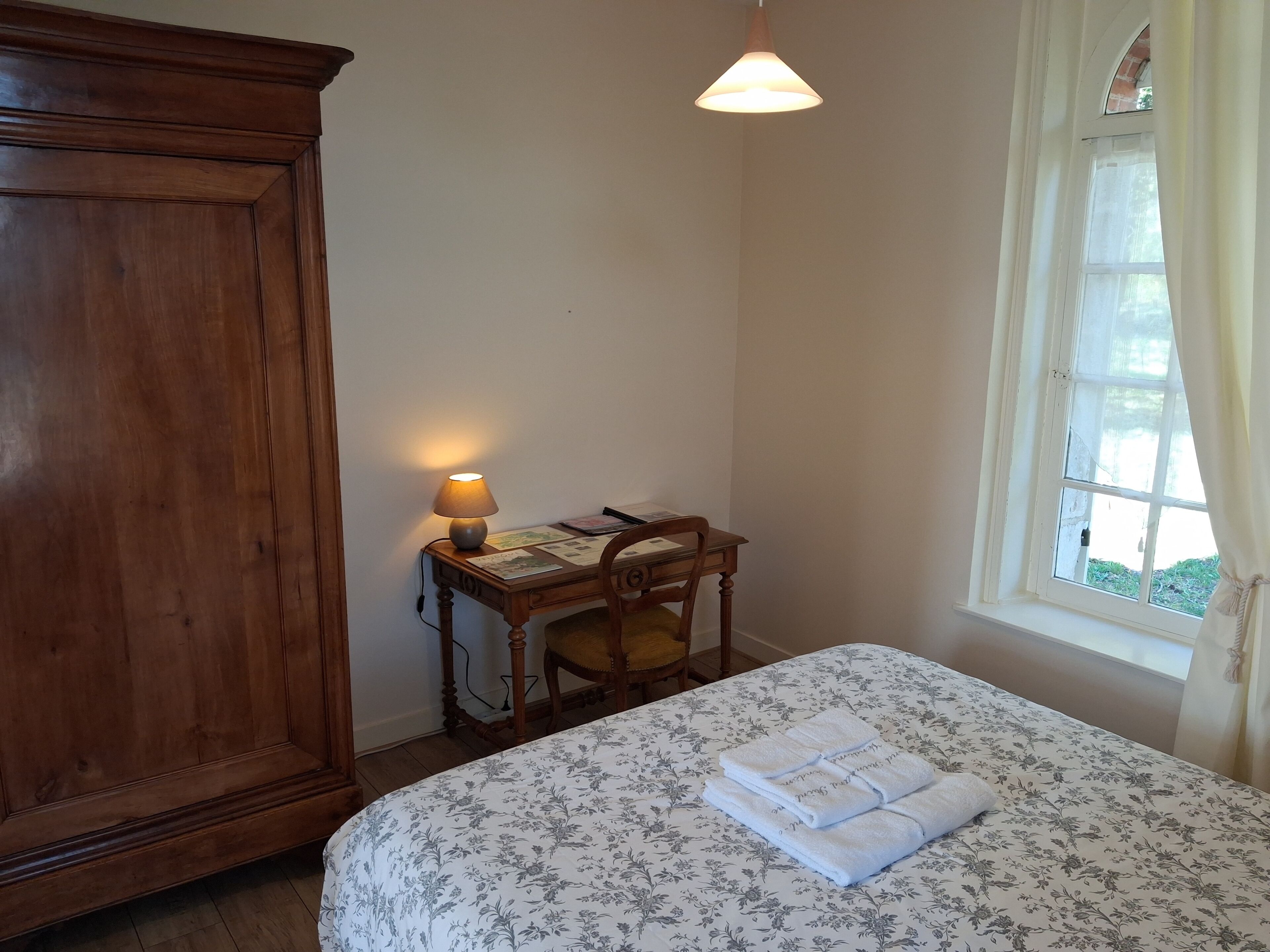 A bedroom with a wooden wardrobe, a desk with a lamp, a bed with a floral bedspread, and a window with curtains.
