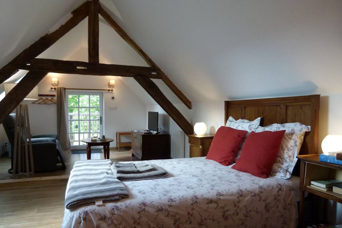 Ein Schlafzimmer mit schrägen Wänden, einem Bett mit floralem Bettbezug und roten Kissen, einem Nachttisch mit Lampe sowie einem Fenster mit Vorhängen.